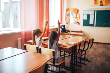 Two little schoolgirls sit at a desk in a school class and raise their hands.