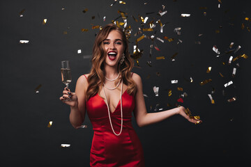Girl in silk dress celebrates, holding champagne on black background with confetti