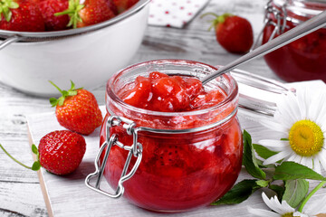 Strawberry jam in glass jar on the white wooden table. Berries around