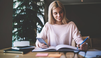 Successful female organisator of knowledge events scribing notes from mobile organiser in textbook for next meeting,happy hipster girl preparing to university test using cellular for browsing internet