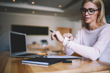 Beautiful woman looking at smartwatch sitting at desk with modern laptop computer and folder, checking time to deadline waiting end of work checking notification on wearable gadget touchscreen