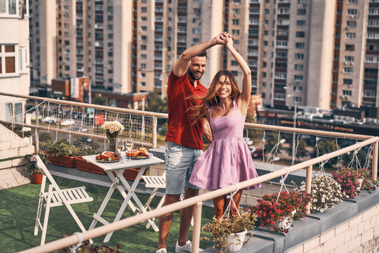 Playful young couple in casual clothing dancing and smiling while having romantic dinner on the rooftop patio outdoors - Powered by Adobe