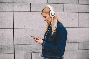 Side view of hipster girl dressed in denim apparel choosing audio songs in music application on smartphone for listening in modern headphones walking along grey promotional background