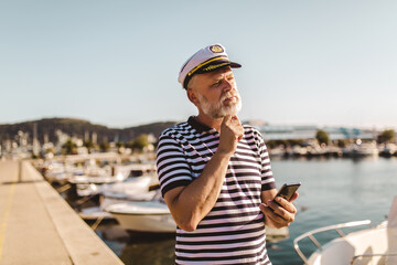 Fototapeta na wymiar Mature man stands on pier dressed in a sailor's shirt and hat using smart phone.