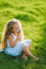 Little blonde girl with long hair has fun eating ice cream in a summer park. Little blonde girl with long hair has fun eating ice cream in a summer park. Sits on the grass with ice cream.