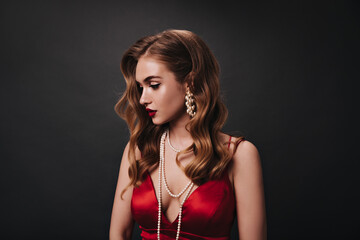 Attractive woman in red top posing on black background. Sad curly lady in satin dress looks down on isolated backdrop