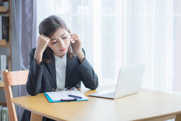 A beautiful Asian woman wearing Thai formal attire, she is sitting at her desk and talking on the phone She touches her head like a headache and looks stressed, as if there is a problem or anxiousness