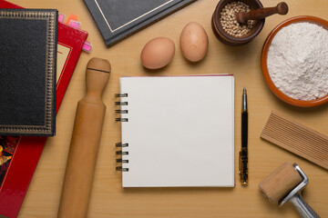 Top view of open blank recipe book next books, eggs, flour and cookware set.