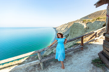 A beautiful young woman in a blue dress sits on a rock on the shore of the azure coast of the sea.