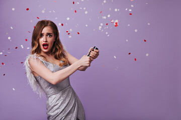 Surprised woman in shiny outfit throwing confetti on purple background. Shocked blonde lady in silver dress has party and posing on isolated backdrop