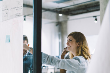 Cheerful caucasian young woman laughing while pointing on plan during teamwork with colleagues in modern office.Positive female coach in formal wear conducting workshop standing behind glass wall