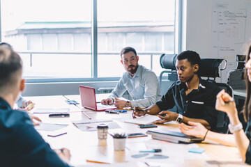 Team of employees dressed in formal wear discussing ideas for new project during briefing at table with laptops in modern office.Multicultural group of young entrepreneurs having business meeting