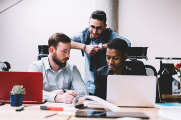 Multicultural team of office employees collaborating on common project at meeting table with laptops.Diverse young people in formal wear analyzing working plan and cooperating on developing startup