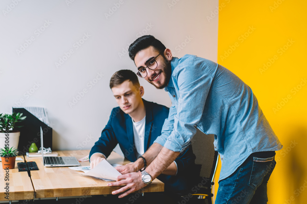 Poster portrait of cheerful young man smiling at camera while teamworking with colleague on developing comm