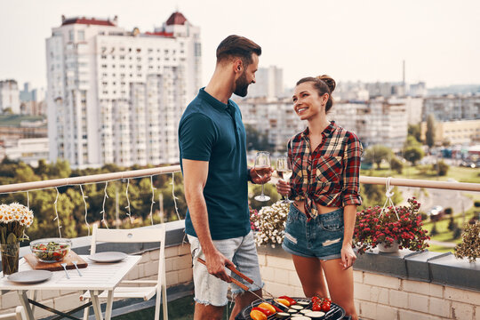 Beautiful young couple in casual clothing preparing barbecue and smiling while standing on the rooftop patio outdoors - Powered by Adobe