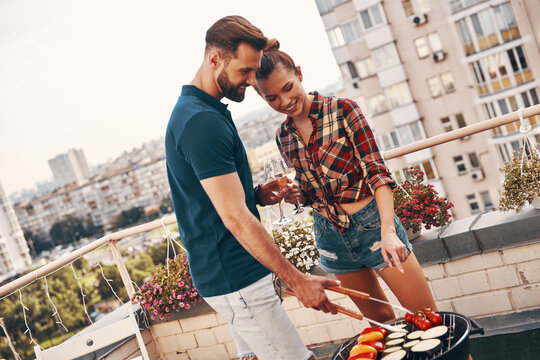 Cute young couple in casual clothing preparing barbecue and smiling while standing on the rooftop patio outdoors