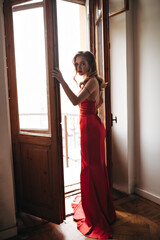 Lady in long dress opening balcony door and looking into camera. Attractive blonde woman in silk red outfit posing against window