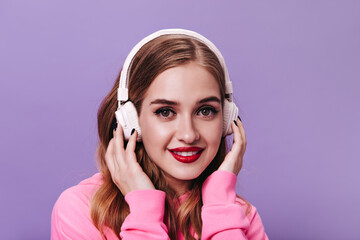 Greeneyed woman in pink hoodie looking into camera and listening to music in headphones. Pretty lady in stylish sweatshirt smiling on purple backgroud