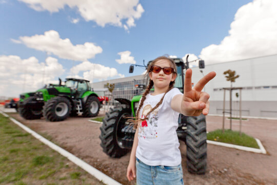 A girl with pigtails in a light T-shirt and jeans poses at the tractor. Day. Summer. Sunny.