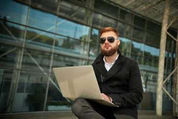 Fototapeta premium Manager holding laptop, Out of office work, young businessman in sunglasses using computer for his job at outside. Handsome bearded hipster, Communication and technology, in the open air, Surfing