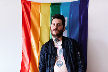 young Caucasian boy with a beard and tattoos poses in front of a rainbow flag of peace with a military jacket.