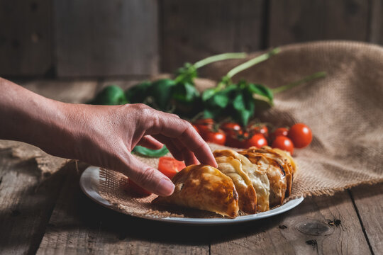 Unrecognizable man picking up a traditional Spanish homemade turnovers served in dish on rustic wooden table with fresh spinach and tomatoes