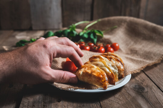 Unrecognizable man picking up a traditional Spanish homemade turnovers served in dish on rustic wooden table with fresh spinach and tomatoes