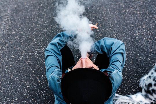 From above view of unrecognizable female in stylish black hat and casual clothes sitting on asphalt road and smoking cigarette while resting during walk