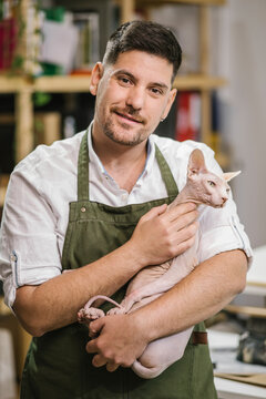 Joyful unshaven artisan in white shirt and green apron smiling at camera while carry calm Sphynx cat on hands in modern studio