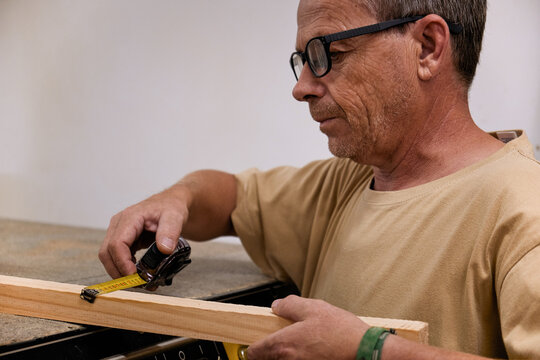 Crop focused carpentry worker in glasses and casual clothes controlling size of wooden detail using measuring tape while working in light modern workshop