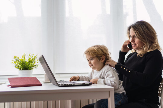 Side view of female remote employee holding curious little child on knees while sitting at table and working with laptop at home