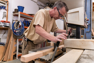 Low angle of attentive middle aged male woodworker in glasses and casual clothes focusing and drilling lumber using special electric machine while working in contemporary workshop