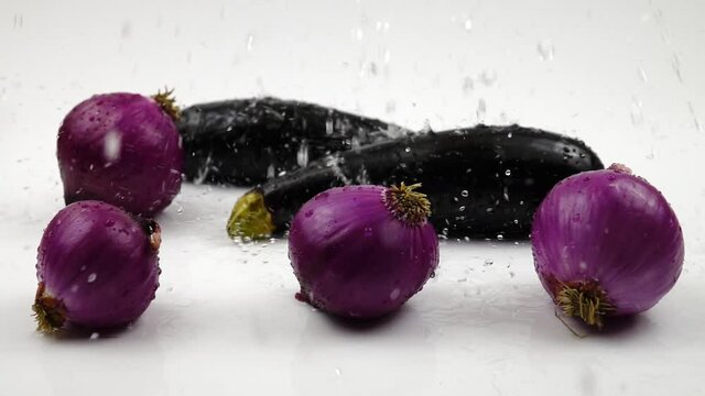 Trickles of water pour onto the red onions and eggplants, lying in the water.  Slow motion, white background