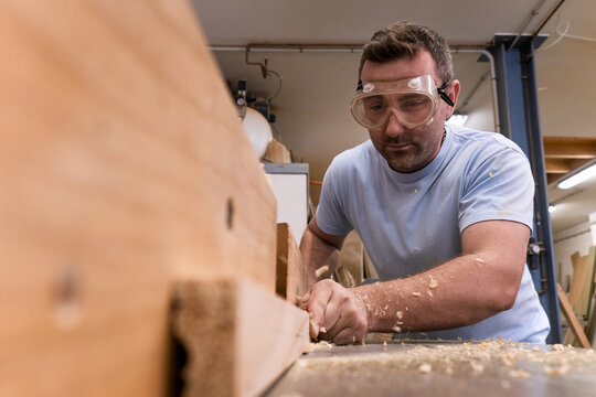 Low angle of focused carpenter in protective glasses milling timber while crafting detail using electric machine in light modern workshop