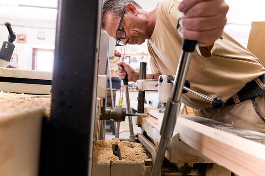 Low angle of attentive middle aged male woodworker in glasses and casual clothes focusing and drilling lumber using special electric machine while working in contemporary workshop