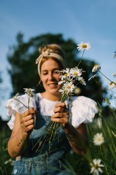 Blurred Young Woman In Casual Clothes Enjoying Smell Of Fresh Chamomiles While Collecting Flowers On Green Meadow In Spring