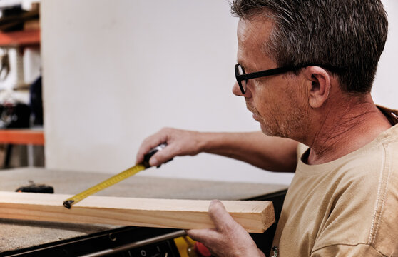 Crop focused carpentry worker in glasses and casual clothes controlling size of wooden detail using measuring tape while working in light modern workshop