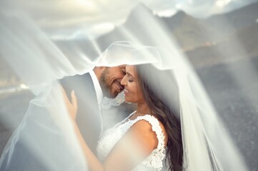 Side view of content bride in wedding gown and groom in suit cuddling with closed eyes and kissing under veil on background of mountains