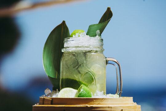 Glass mug with refreshing cold cocktail with fresh lime and green leaves composed against blurred background