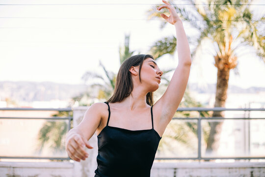 Graceful female dancer in moment of performing element with outstretches arms and closed eyes on summer terrace on background of palm trees