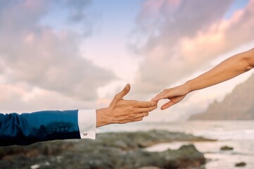 Unrecognizable groom in elegant suit and bride gently touching fingers while standing on rocky coast during magnificent sunset