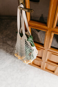 Reusable white string bag with fresh loquat fruits on branches hanging on knob of wooden door inside room with dark light