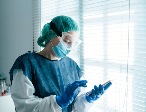 Side view of female medical specialist in protective gown with mask and goggles browsing on mobile phone and looking out shuttered window while working in hospital during coronavirus outbreak