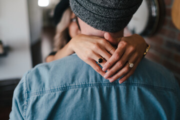 Back view of unrecognizable boyfriend in denim jacket and sock cap embracing blond girlfriend with rings on hands while standing together at home