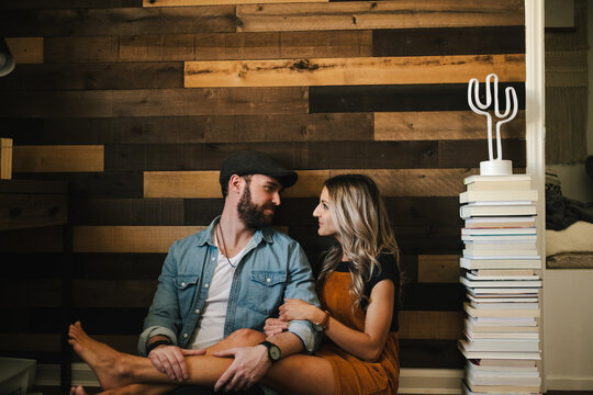 Smiling bearded boyfriend in cap and denim jacket cuddling cheerful blond girlfriend while sitting near wooden wall and looking at each other with love
