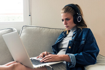 Focused young female freelancer in casual wear with wireless headphones sitting on sofa and using laptop while working on remote project at home