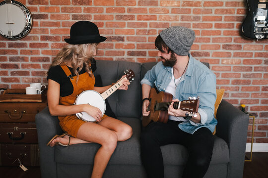 Smiling woman sitting with crossed legs on sofa and playing banjo near boyfriend in denim jacket and sock cap playing acoustic guitar on weekend