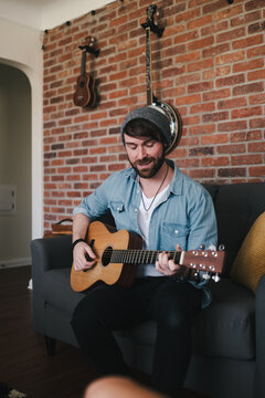 Cheerful bearded guitarist in sock cap and denim jacket playing acoustic guitar while sitting on couch with decorative pillow near brick wall with different musical instruments at home