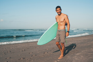 Happy shirtless ethnic male in shirts with blue surfboard looking away while standing on sandy beach near sea in sunny day