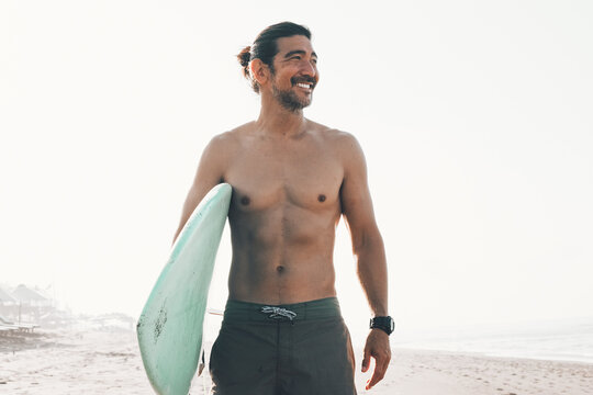 Happy shirtless ethnic male in shirts with blue surfboard looking away while standing on sandy beach near sea in sunny day